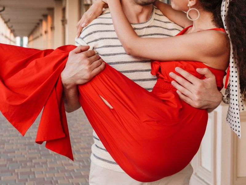 Ein Bild von einem jungen Mann, der seine Frau, die ein rotes Kleid trägt, auf Händen trägt.