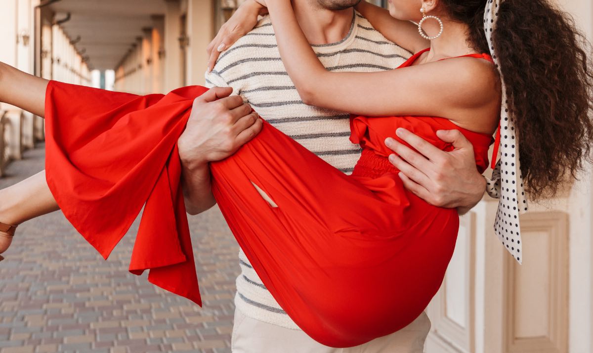 Ein Bild von einem jungen Mann, der seine Frau, die ein rotes Kleid trägt, auf Händen trägt.