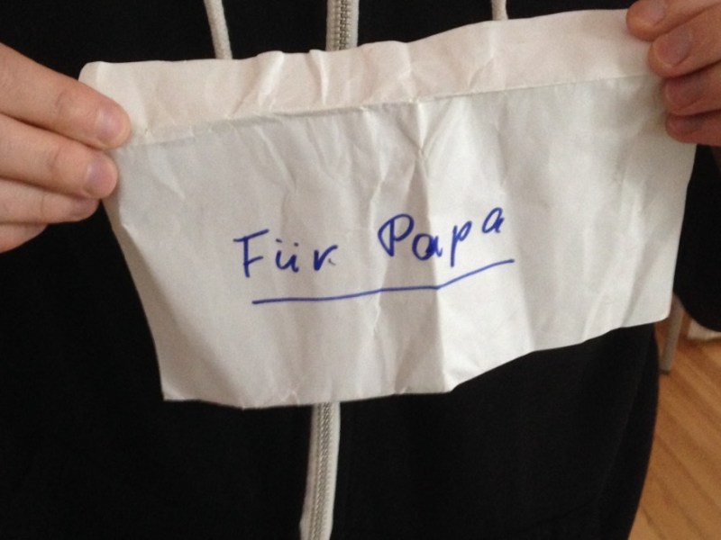 Ein Mann mit Ehering am Finger hält einen zerknitterten Briefumschlag hoch, auf dem 'für Papa' steht.