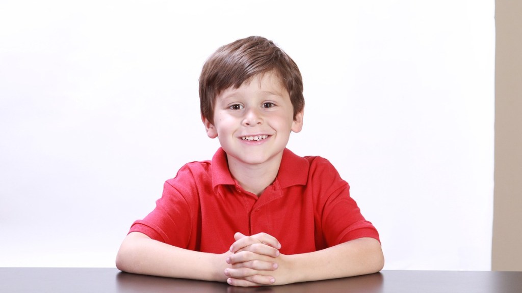 Ein Junge mit rotem Hemd sitzt an einem Tisch und lächelt.