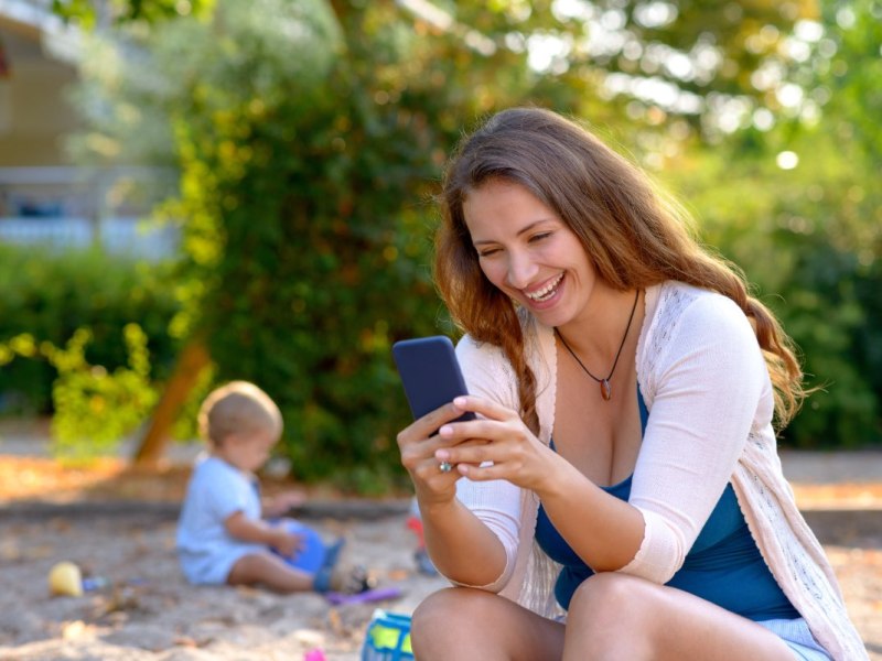 Eine Mutter sitzt auf einem Spielplatz und schaut auf ihr Handy.
