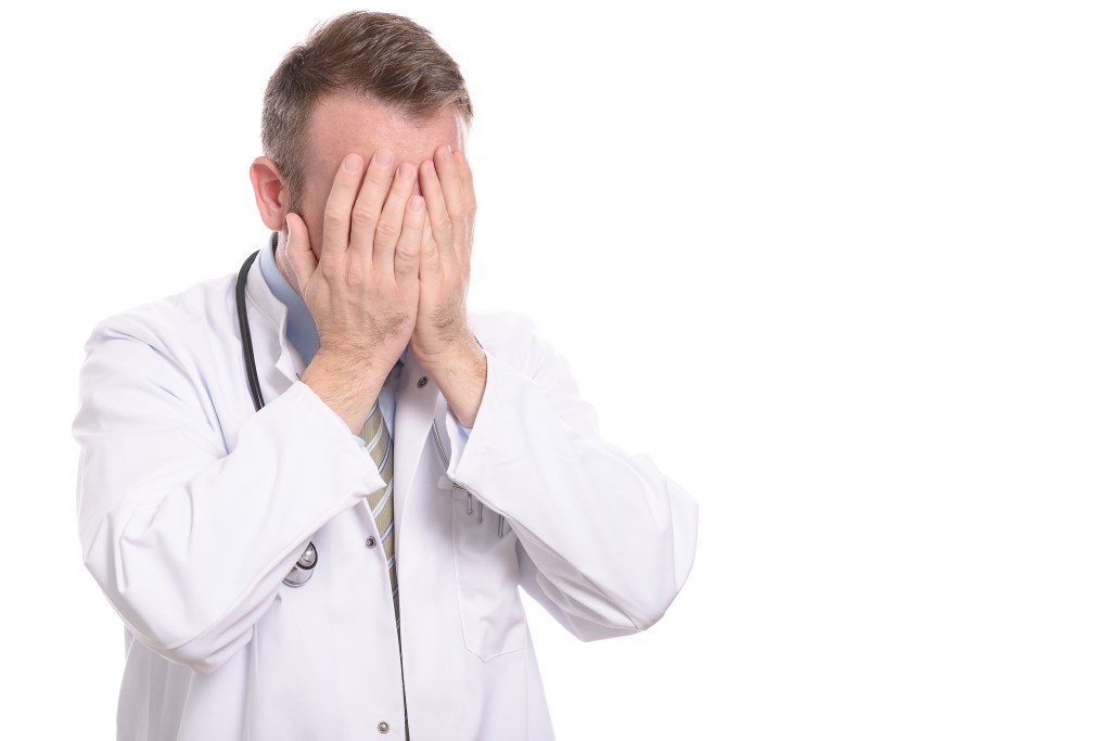 Ein Arzt mit Stethoskop und weißem Kittel schlägt verzweifelt die Hände vors Gesicht.