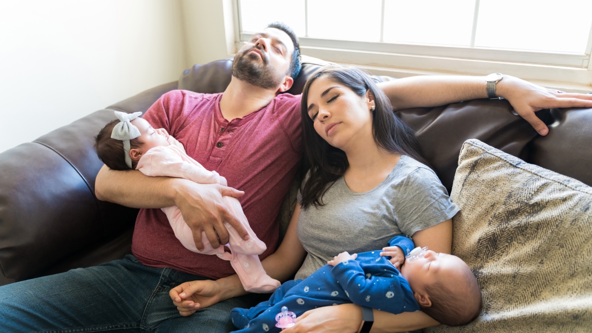Zwei schlafende Eltern liegen auf einem Sofa und halten ihre Babys im Arm.