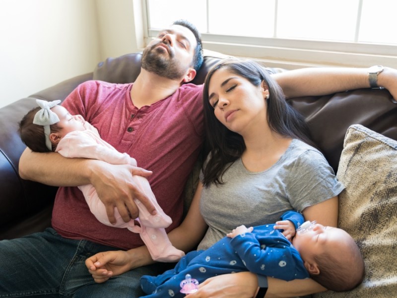Zwei schlafende Eltern liegen auf einem Sofa und halten ihre Babys im Arm.