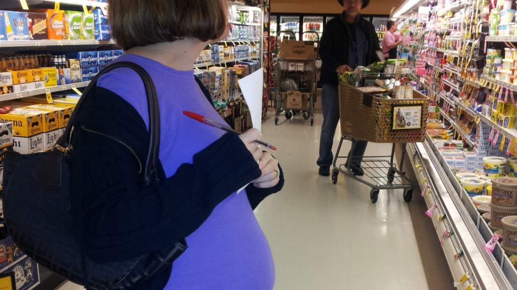 Eine schwangere Frau steht mit Einkaufszettel in der Hand in einem Supermarkt.