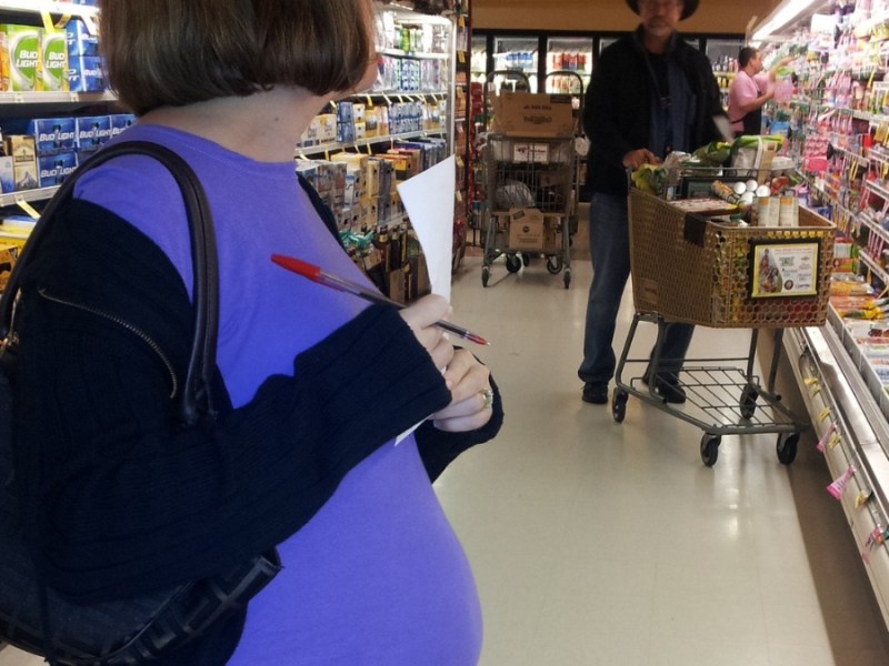 Eine schwangere Frau steht mit Einkaufszettel in der Hand in einem Supermarkt.