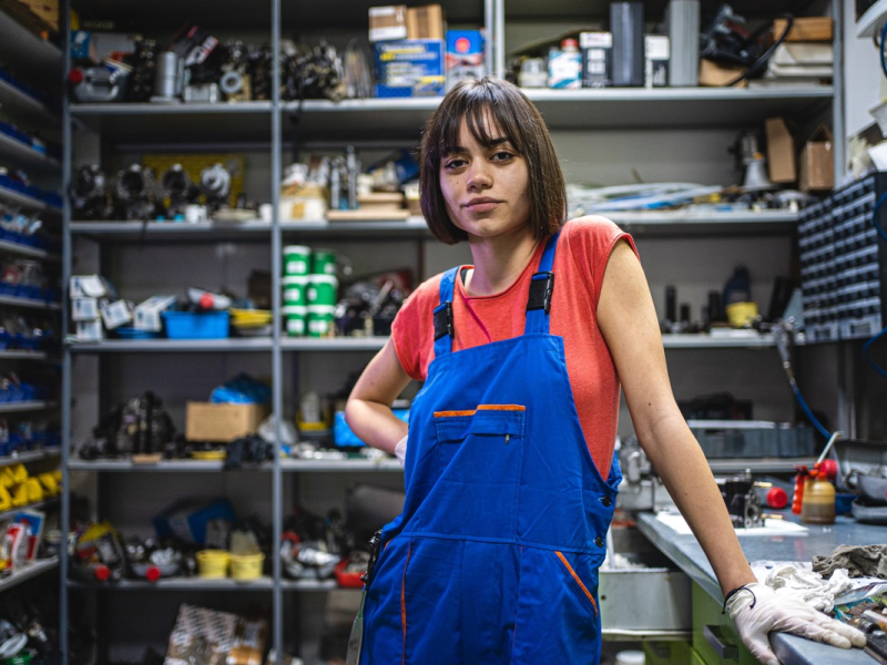 Eine junge Mechanikerin mit blauer Latzhose steht in ihrer Werkstatt.