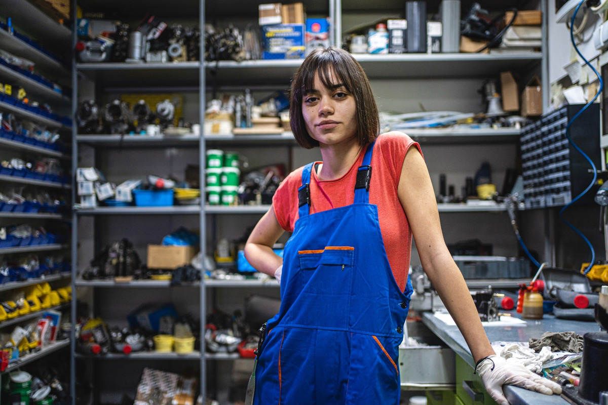 Eine junge Mechanikerin mit blauer Latzhose steht in ihrer Werkstatt.