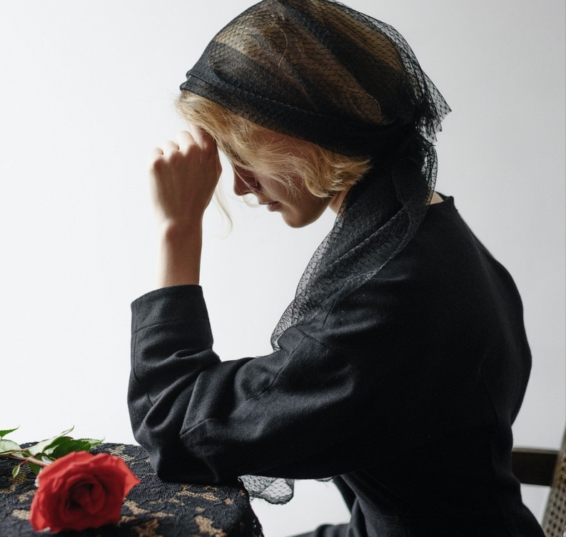 Eine trauernde Witwe mit schwarzem Schleier sitzt an einem Tisch, auf dem eine rote Rose liegt.