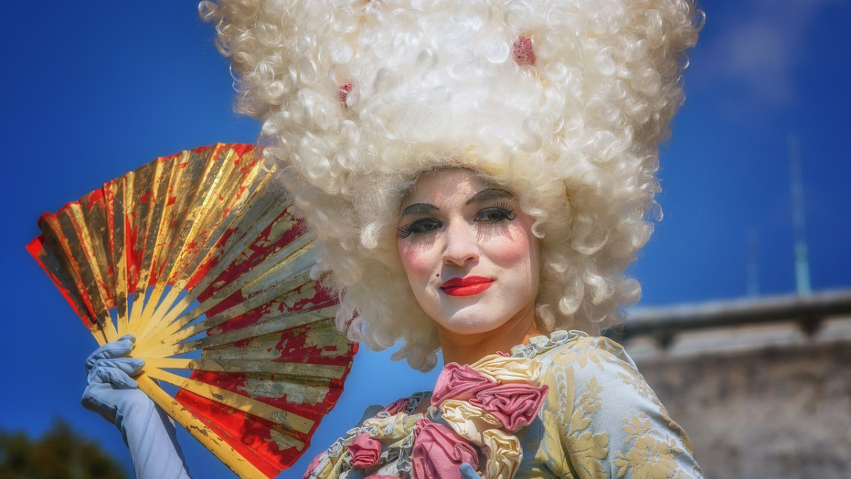 Eine geschminkte Frau in einem barocken Kleid und mit einer Turmfrisur-Perücke, die einen Fächer in der Hand hält.