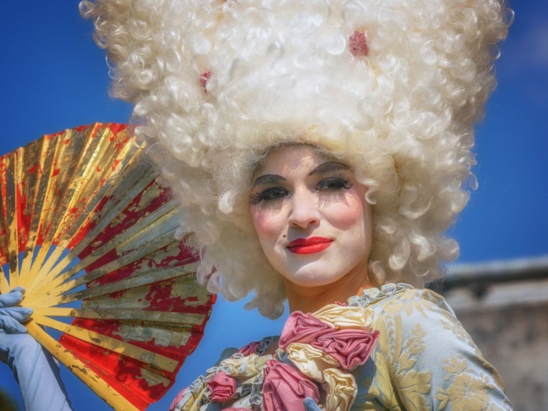 Eine geschminkte Frau in einem barocken Kleid und mit einer Turmfrisur-Perücke, die einen Fächer in der Hand hält.