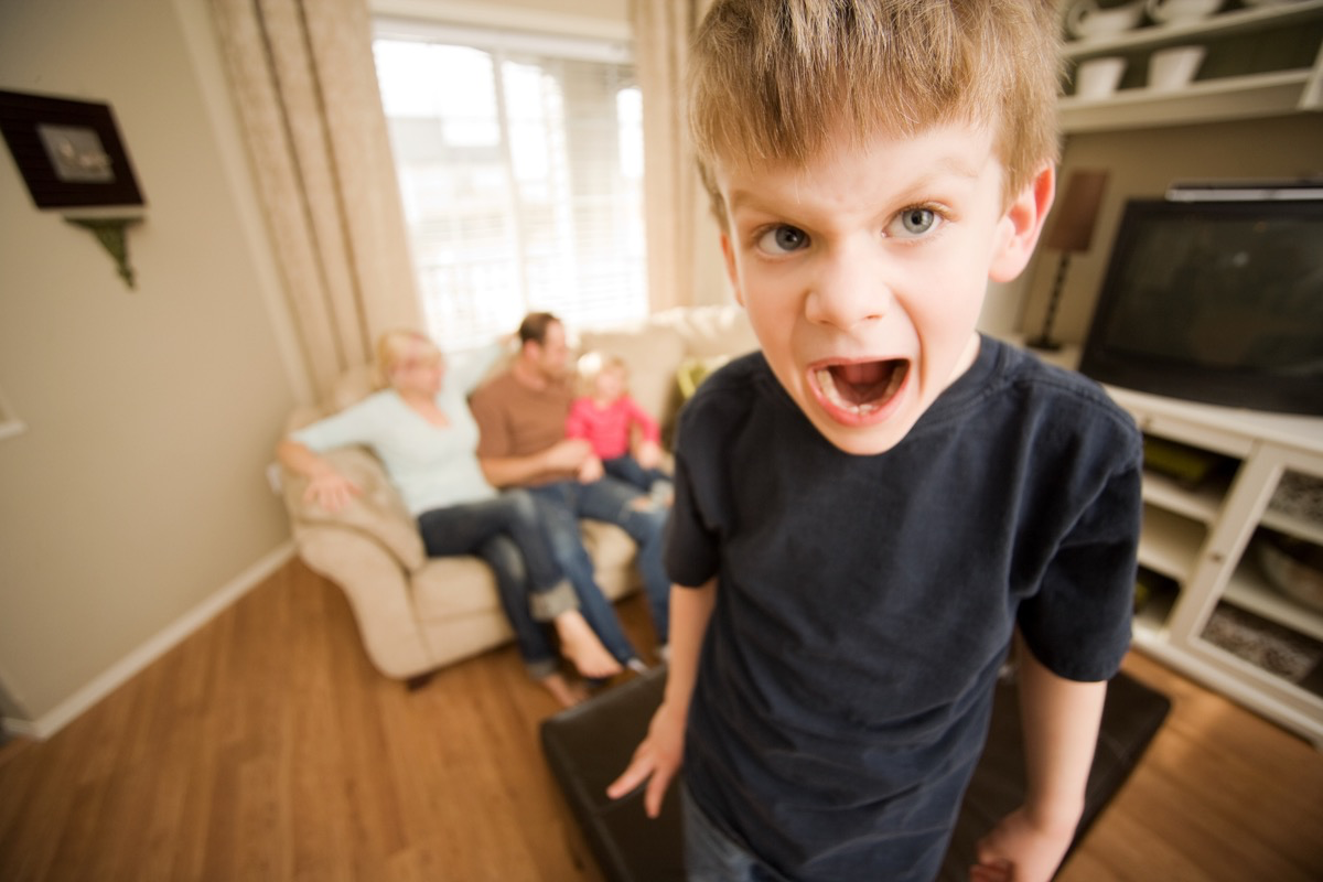 Ein kleiner, blonder Junge steht im Wohnzimmer und schreit, während seine Eltern mit der Tochter auf dem Sofa sitzen