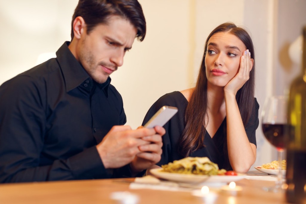 Eine Frau sitzt bei einem Date gelangweilt am Tisch, während ihr Partner auf sein Smartphone schaut.