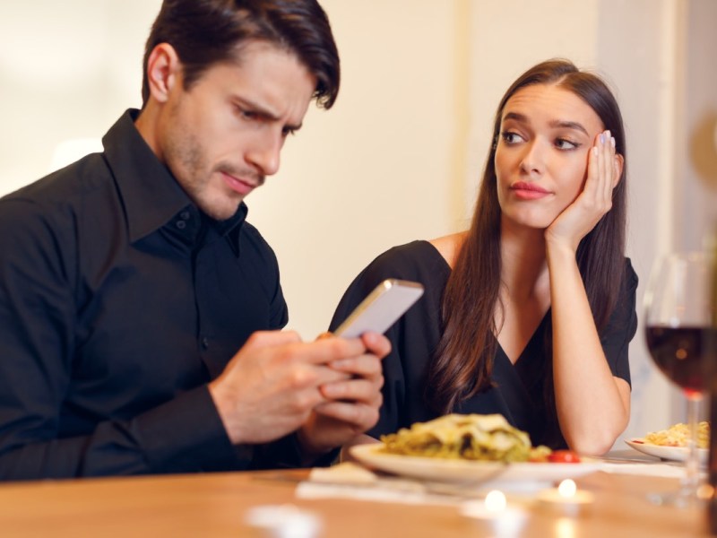 Eine Frau sitzt bei einem Date gelangweilt am Tisch, während ihr Partner auf sein Smartphone schaut.