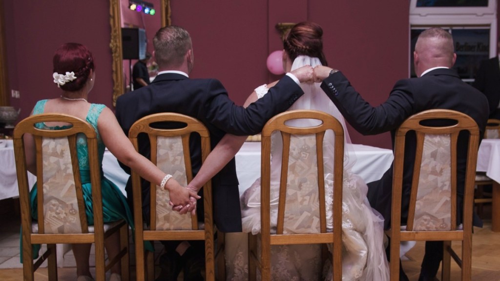 Ein Hochzeitspaar sitzt mit Freunden am Tisch und gibt sich hinter dem Rücken die Hand.