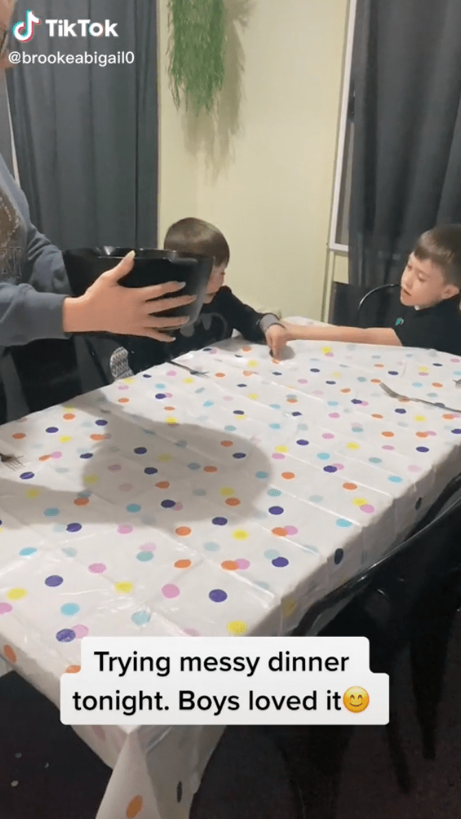 Eine Mutter hält einen Topf mit Spaghetti in der Hand. Ihre Kinder sitzen am Tisch.