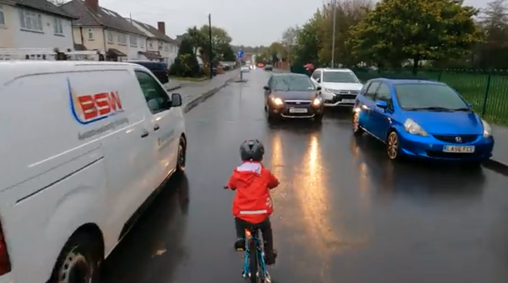 Ein kleiner Junge auf einem Fahrrad mit Fahrradhelm fährt mittig auf der Straße und ein Auto kommt ihm entgegen.