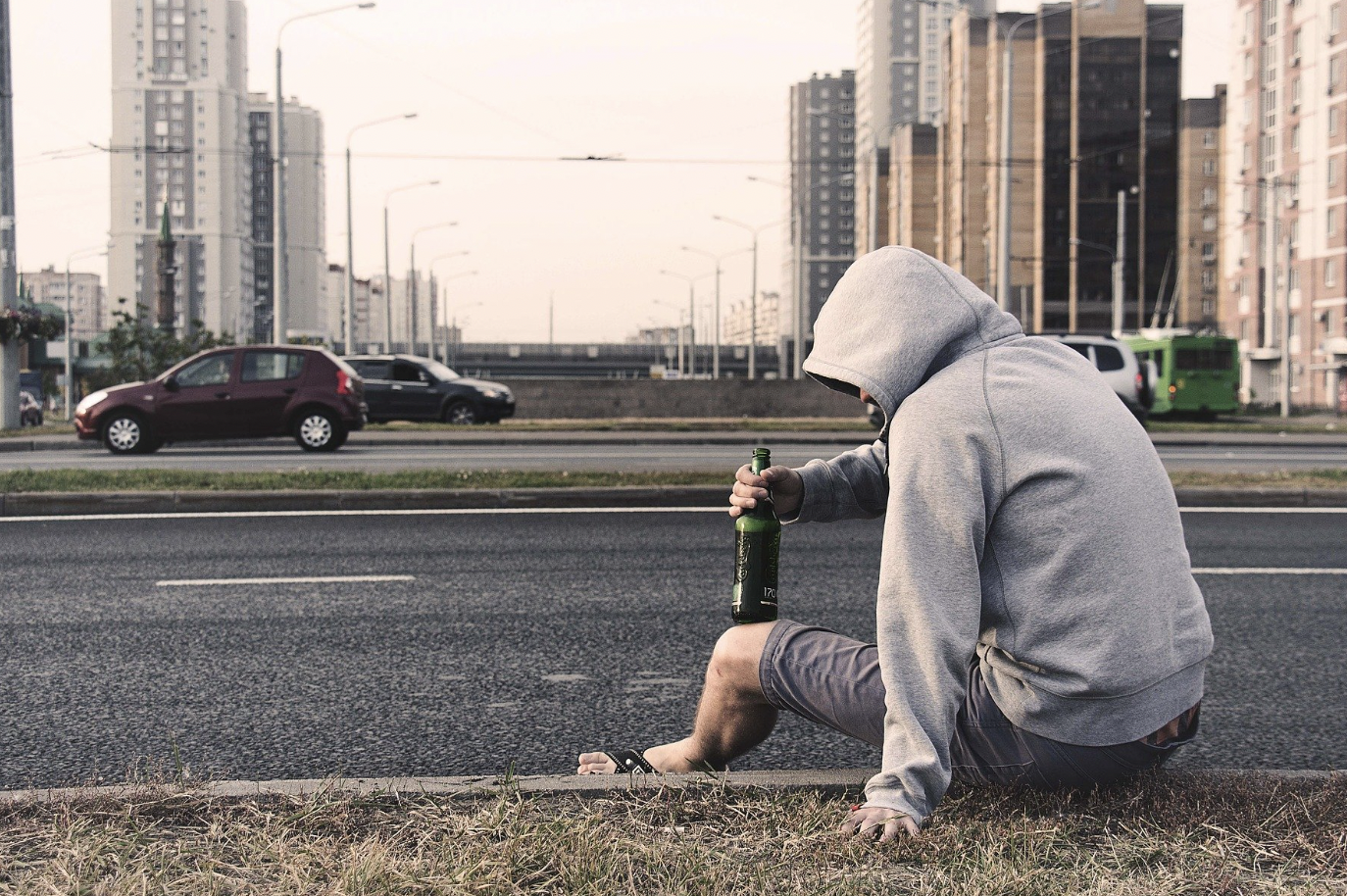 Mann mit Kapuze über Kopf sitzt mit Bierflasche am Straßenrand