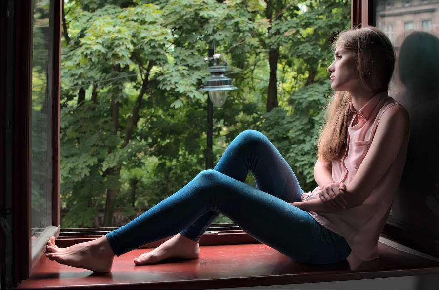Eine blonde Frau sitzt am offenen Fenster auf dem Fensterbrett und schaut gedankenverloren hinaus in den Wald.