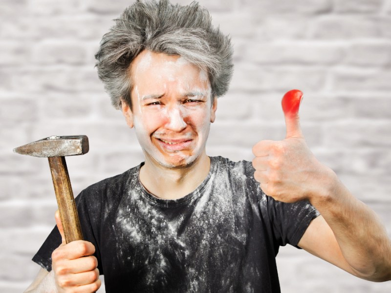 Ein schmutziger Handwerker mit einem Hammer in der Hand hält einen geschwollen Daumen hoch und macht ein schmerzverzerrtes Gesicht.