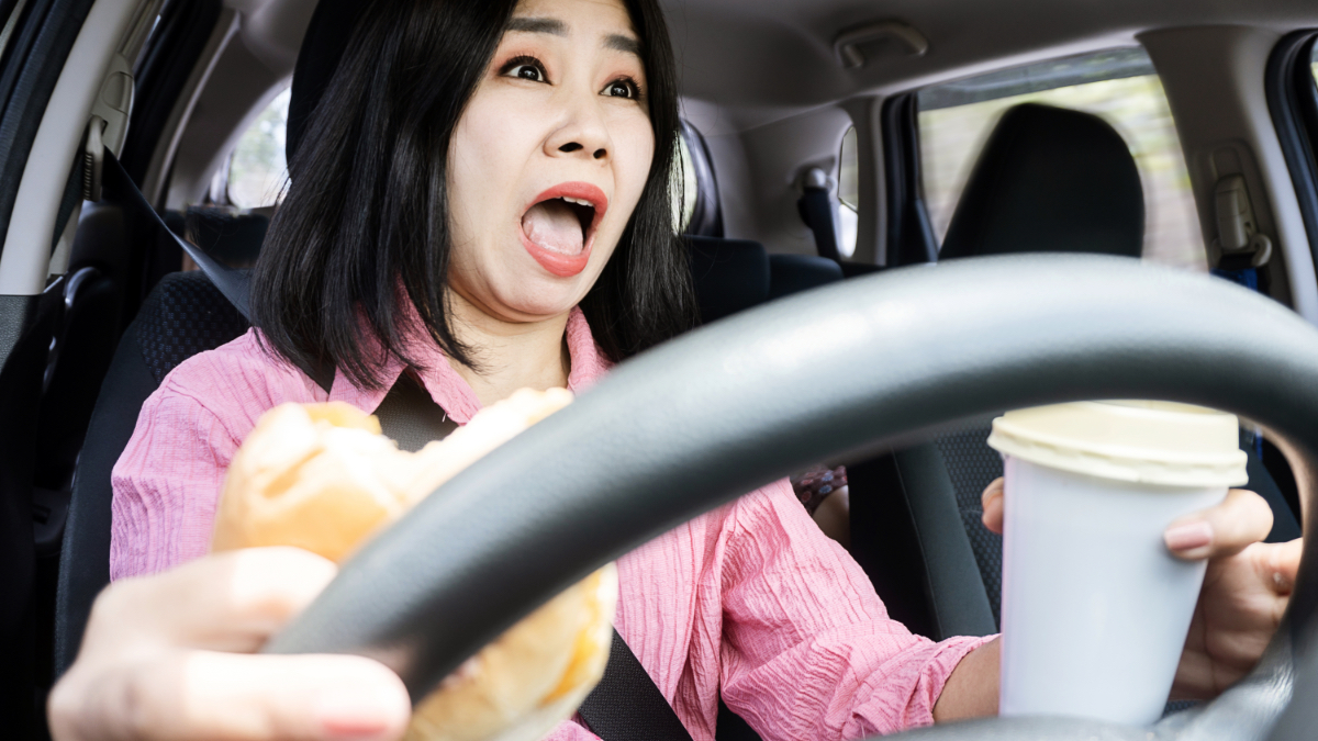 Eine Frau sitzt im Auto und hält mit Getränk und Hamburger in der Hand das Steuer