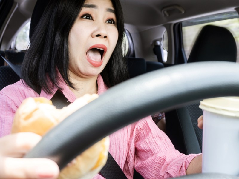 Eine Frau sitzt im Auto und hält mit Getränk und Hamburger in der Hand das Steuer