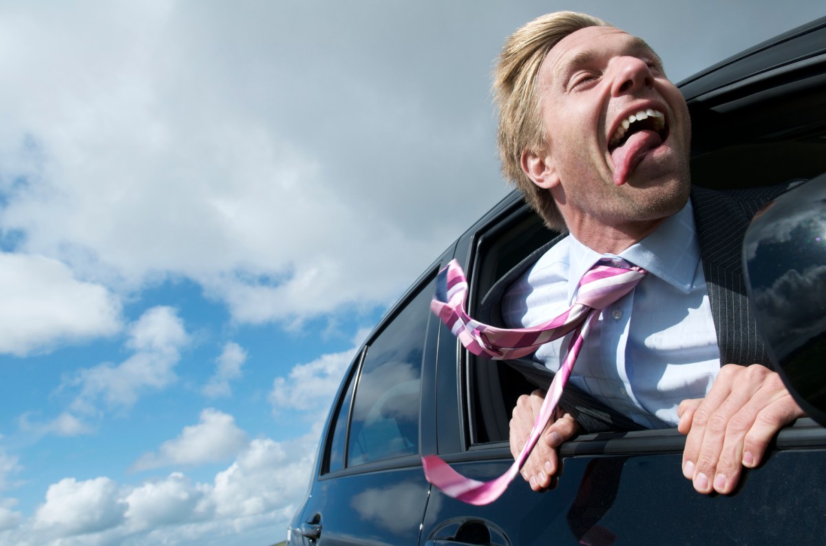 Ein Mann lehnt sich aus dem Fenster eines fahrenden Autos und streckt seine Zunge heraus wie ein Hund.
