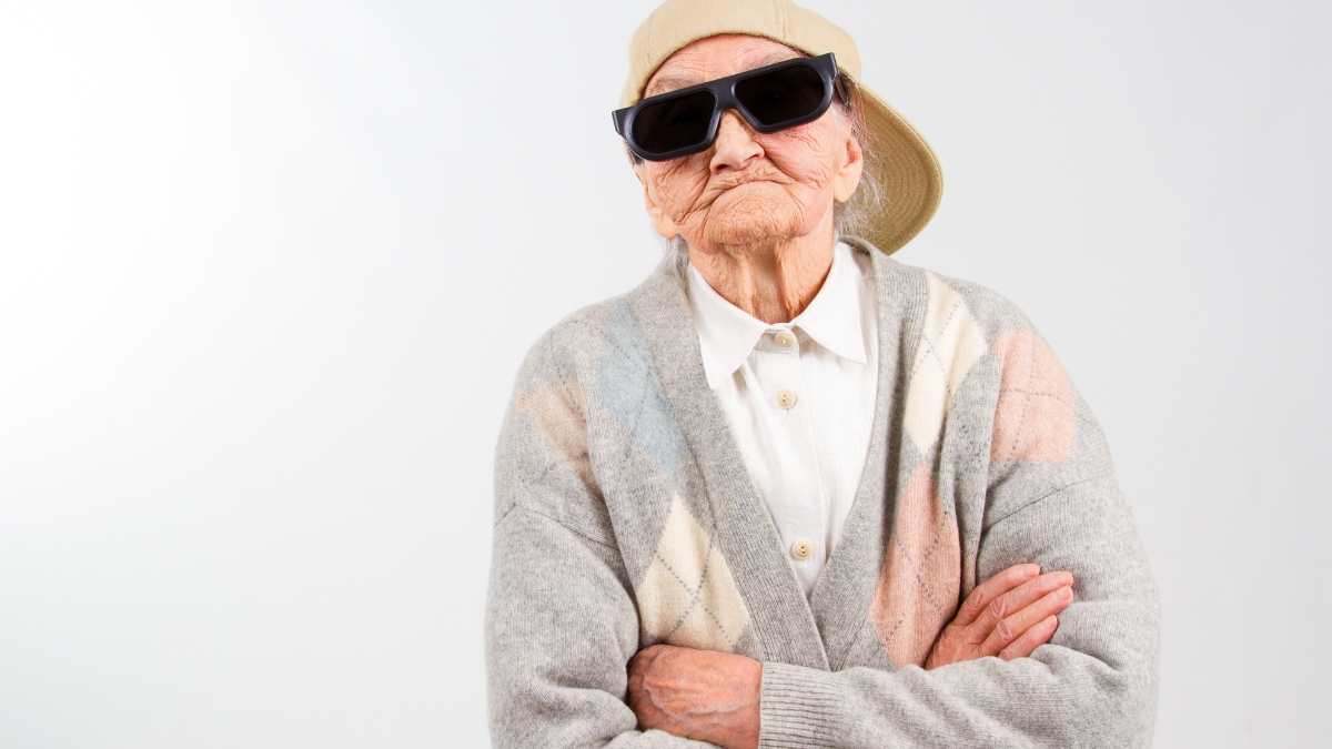 Ein lustiges Porträt einer Rentnerin die mit Schirmmütze, Sonnenbrille und verschränkten Armen posiert.