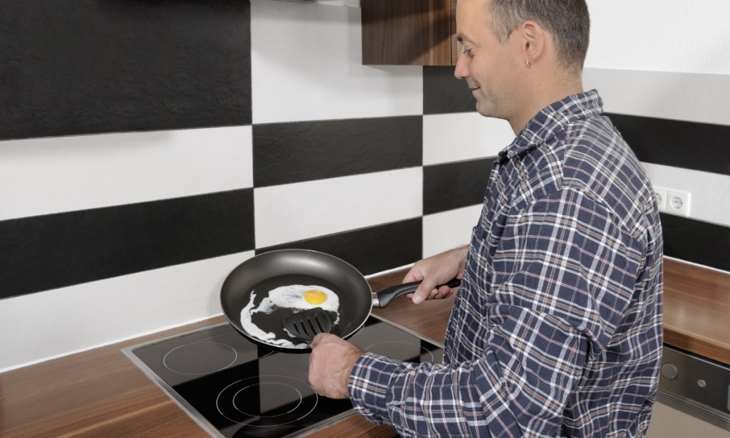 Ein Mann steht in einer Küche am Herd und brät Eier in einer Pfanne.