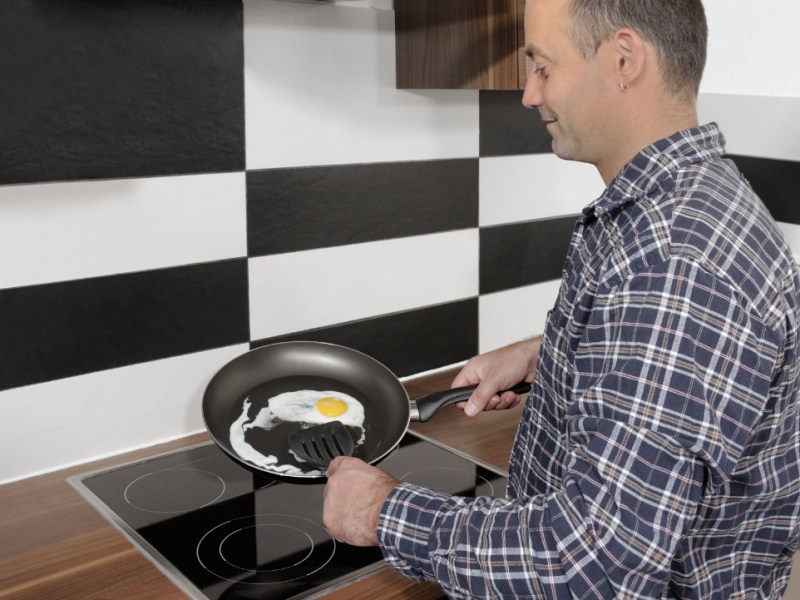 Ein Mann steht in einer Küche am Herd und brät Eier in einer Pfanne.