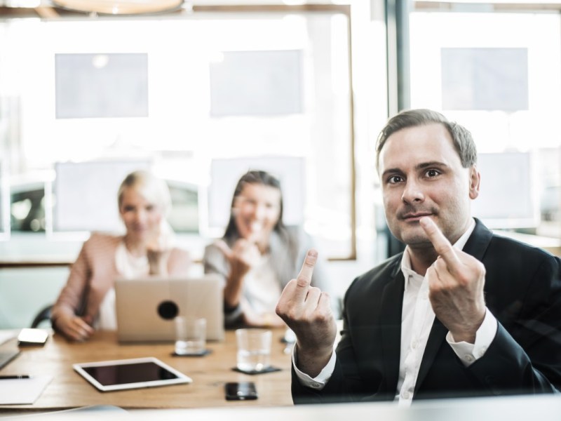 Ein Mann und zwei Frauen in einem Büro lächeln und zeigen der Kamera den Mittelfinger.