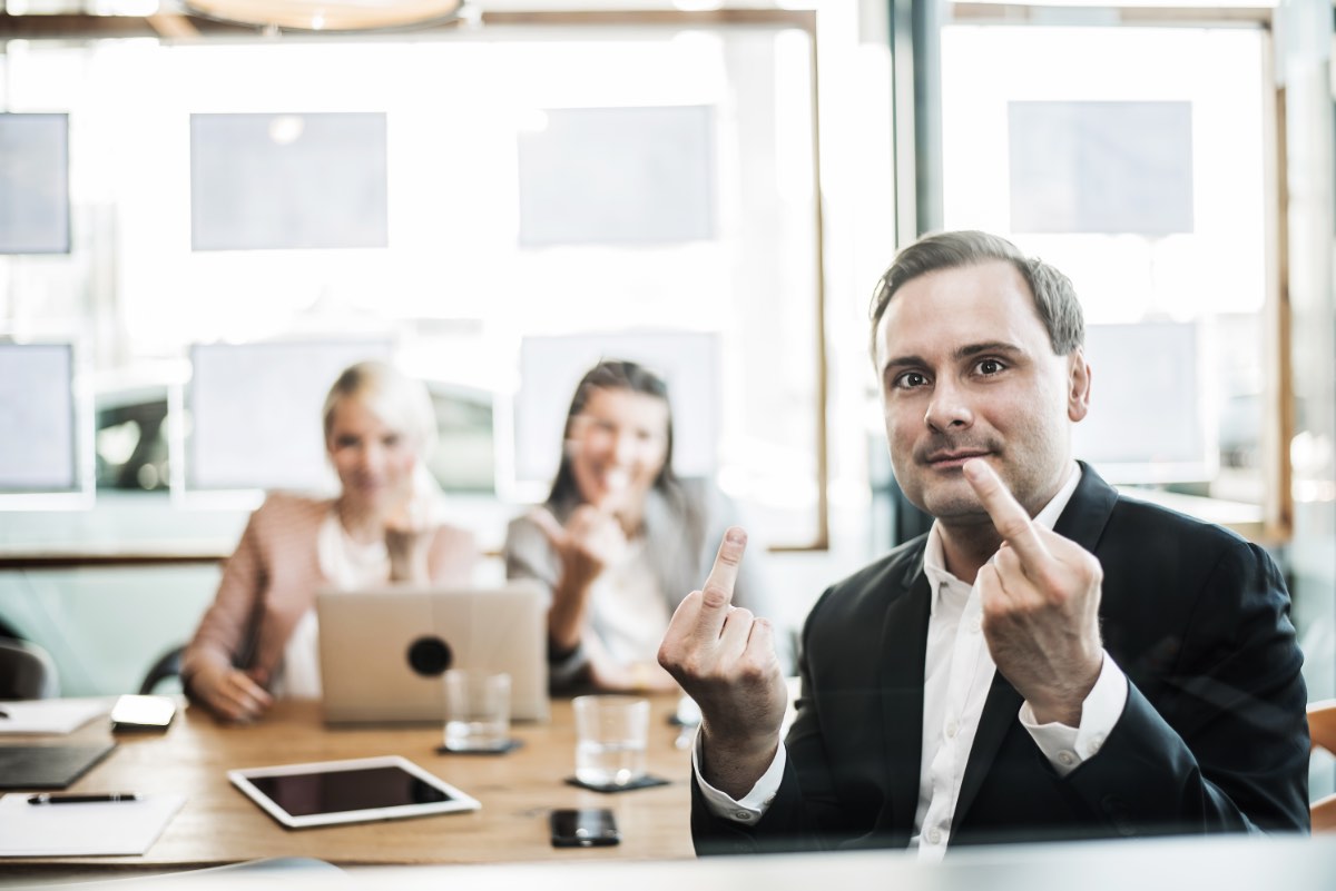 Ein Mann und zwei Frauen in einem Büro lächeln und zeigen der Kamera den Mittelfinger.