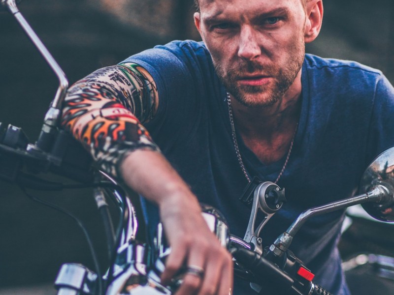 Ein Biker mit Tätowierungen auf dem Arm lehnt sich auf sein Motorrad.