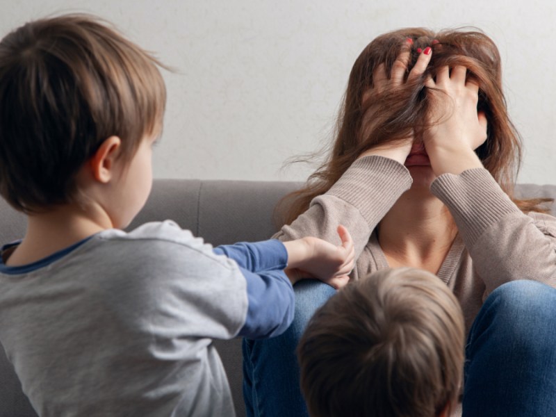 Eine Mutter wird von ihren Kindern genervt und hält sich gestresst die Hände vor das Gesicht.