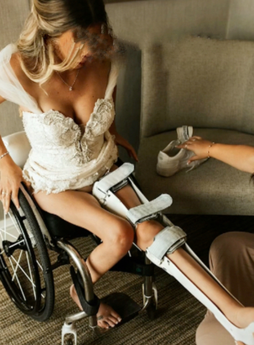 Eine Frau im Brautkleid und geschientem Bein sitzt im Rollstuhl.