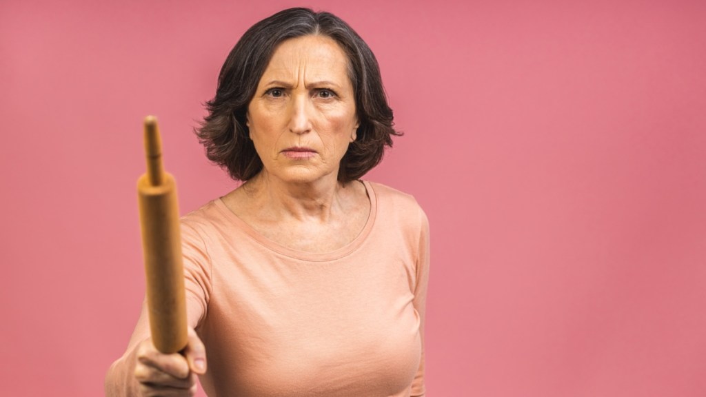 Eine sehr wütende ältere Dame im reifen Alter hält ein Nudelholz und droht, jemanden damit zu schlagen. Isoliert auf rosa Hintergrund.