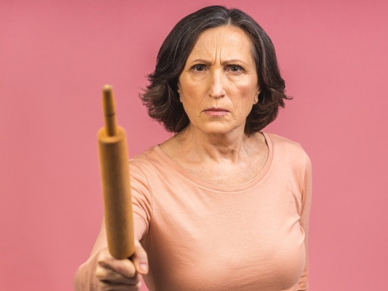 Eine sehr wütende ältere Dame im reifen Alter hält ein Nudelholz und droht, jemanden damit zu schlagen. Isoliert auf rosa Hintergrund.