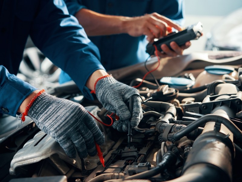Nahaufnahme eines Mechanikers mit Schutzhandschuhen bei der Verwendung eines Multimeters zur Prüfung einer Autobatterie.