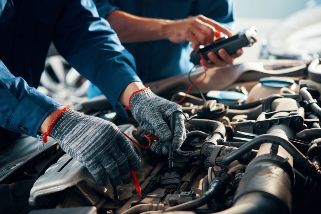 Nahaufnahme eines Mechanikers mit Schutzhandschuhen bei der Verwendung eines Multimeters zur Prüfung einer Autobatterie.