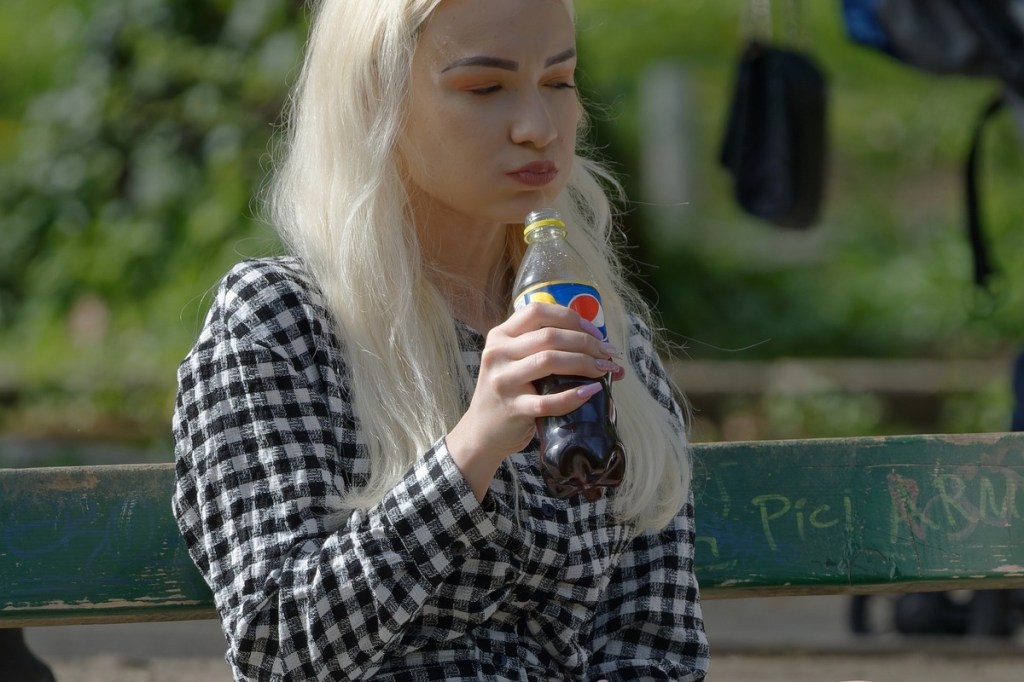 Eine blonde Frau trinkt Pepsi auf einer Parkbank.