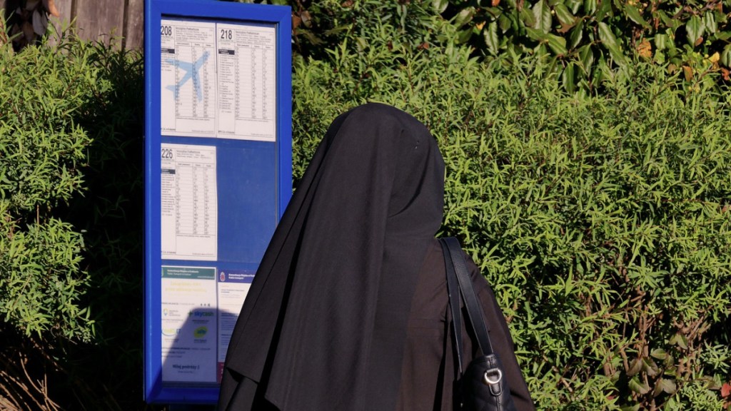 Eine Nonne steht an einer Bushaltestelle vor einem Fahrplan.