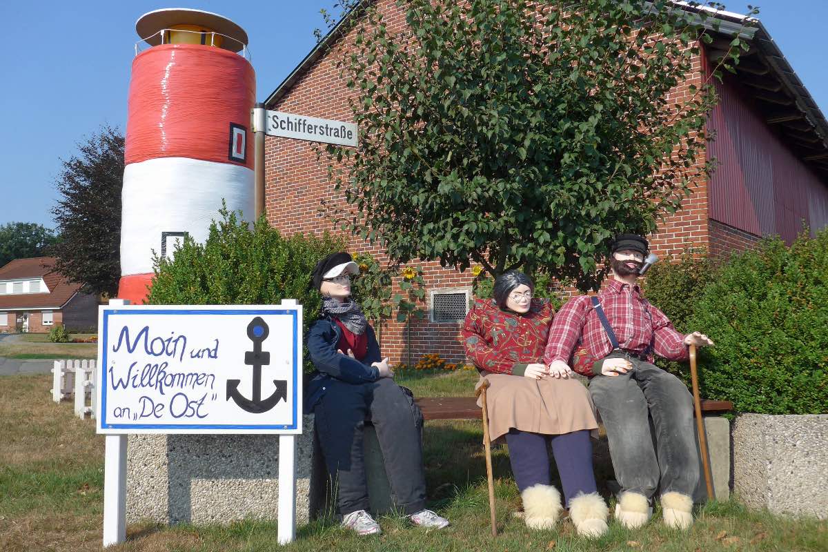 Drei Puppen sitzen auf einer Bank vor einem Leuchtturm in Ostfriesland.