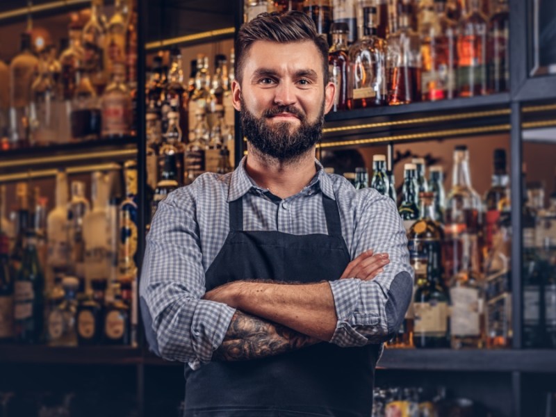 Stilvolle Barkeeper in Hemd und Schürze steht mit verschränkten Armen an der Theke.