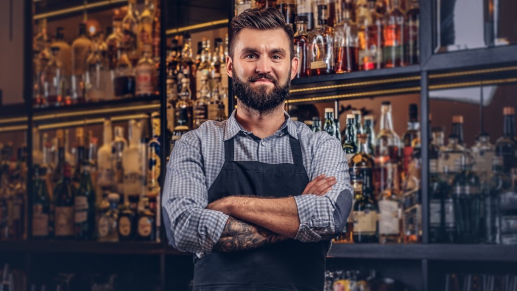 Stilvolle Barkeeper in Hemd und Schürze steht mit verschränkten Armen an der Theke.