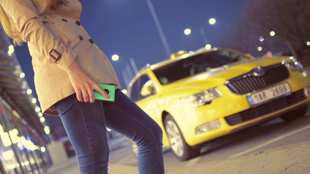 Eine Frau in einem braunen Mantel mit Handy in der Hand wartet abends auf ein Taxi.