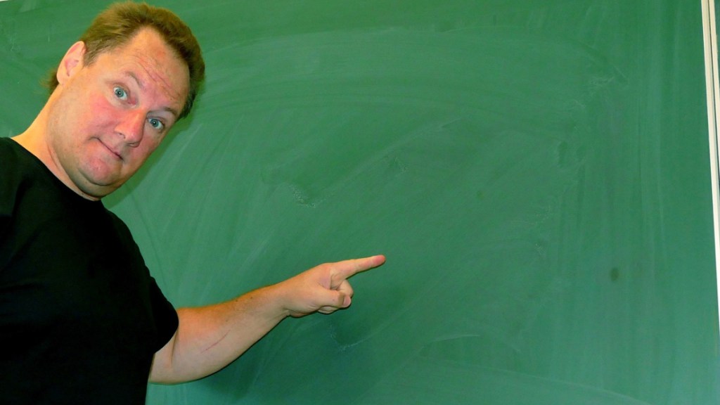 Ein Lehrer zeigt im Klassenzimmer mit dem Finger auf eine leere Tafel.