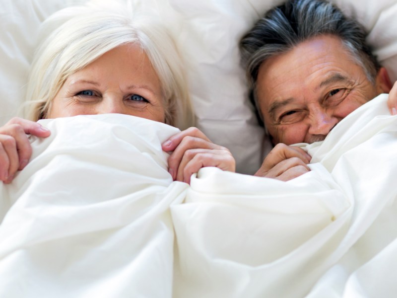 Ein Rentner-Paar liegt im Bett und zieht die Decke über das Gesicht.