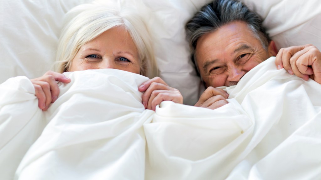 Ein Rentner-Paar liegt im Bett und zieht die Decke über das Gesicht.