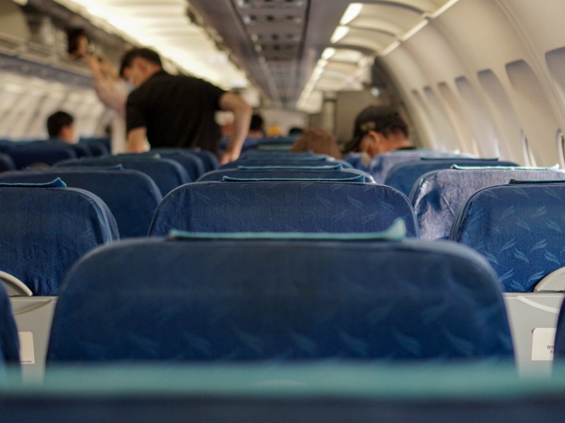 Eine Flugzeugkabine vom Sitz aus fotografiert. Einige Passagiere stehen von ihren Sitzen auf und holen ihr Gepäck aus der Gepäckablage des Flugzeugs.