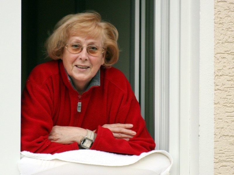 Eine alte Frau mit rotem Pullover schaut aus einem Fenster und stützt sich dabei auf einem Kissen ab.